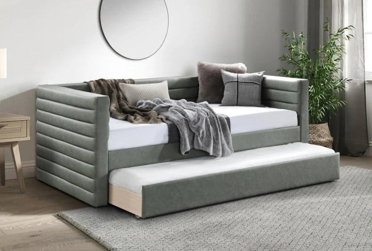 Gray upholstered bed with pull-out trundle in a room setting with a mirror and plant.